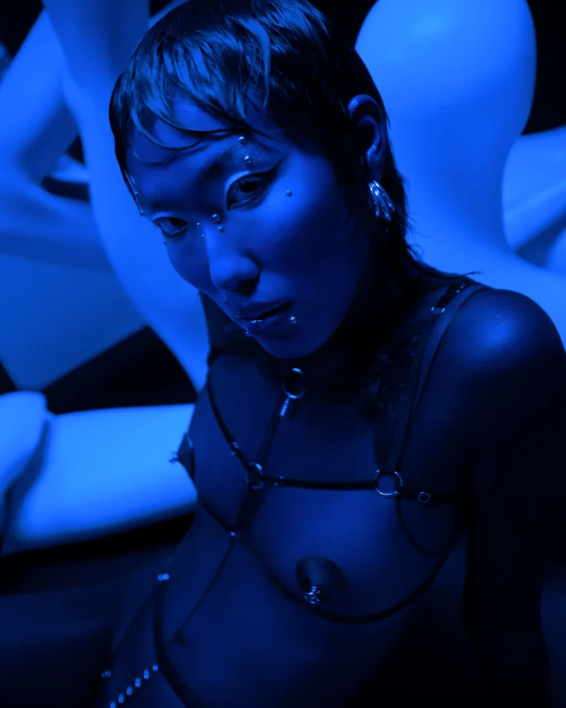 Avant-garde fashion portrait with dramatic blue lighting, face gems and body chain harness