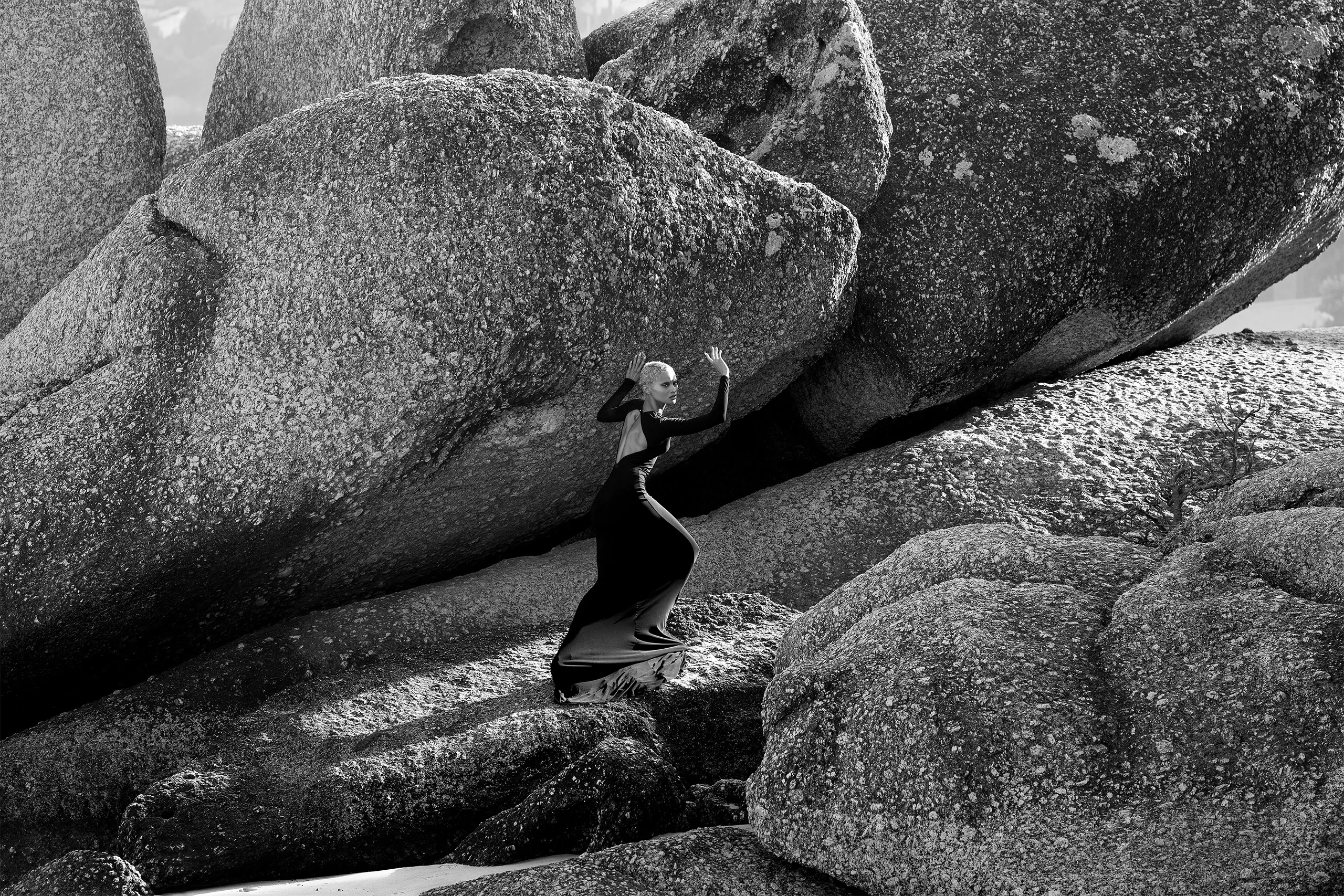 Woman in elegant black gown posing dramatically against massive granite boulders, high fashion editorial