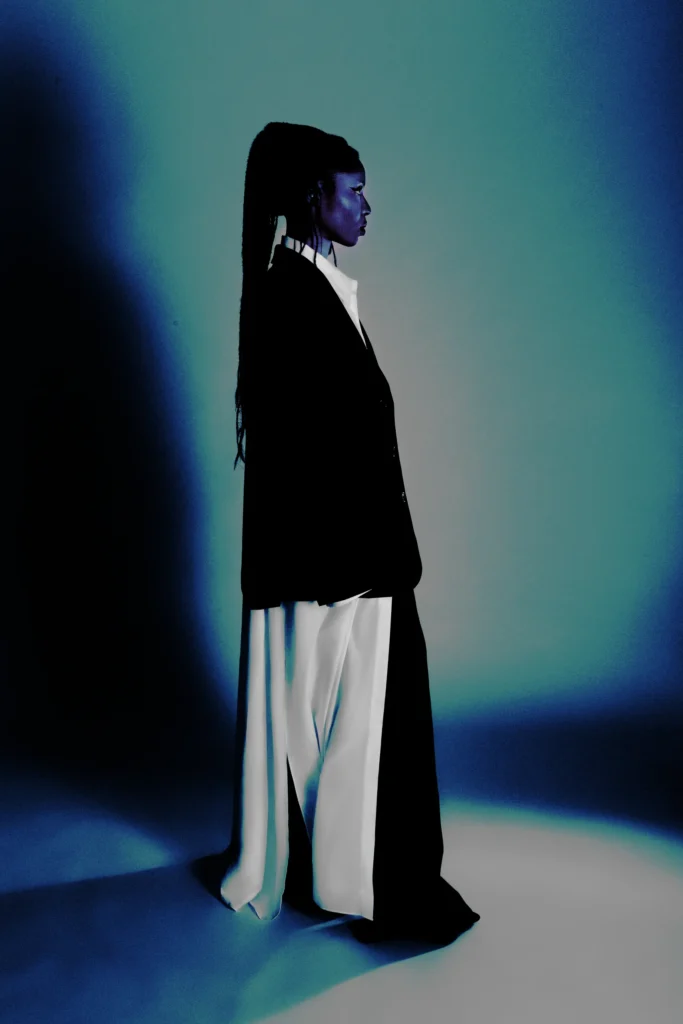 Model in black blazer and white flowing pants, dramatic blue lighting, high fashion studio portrait