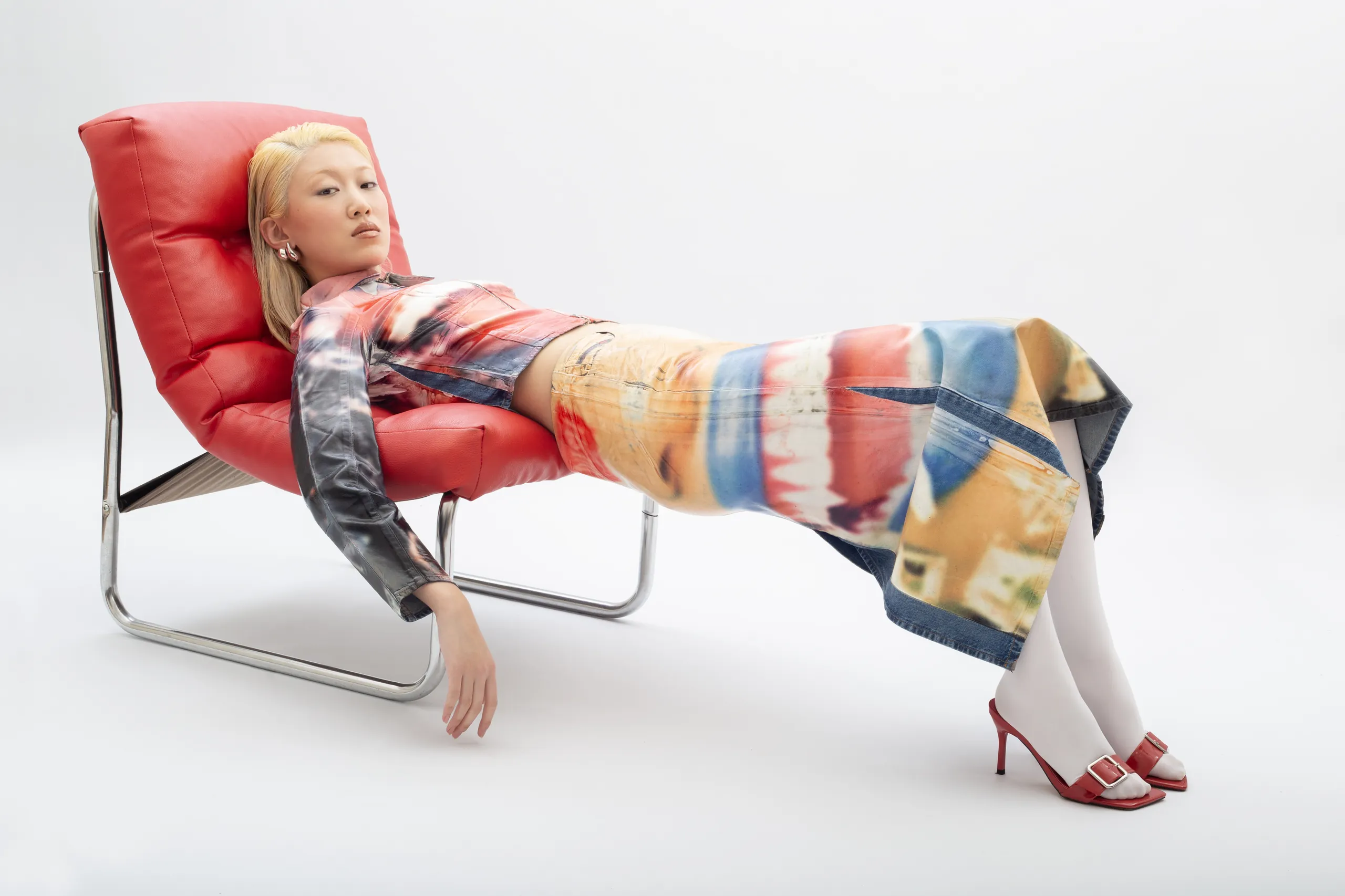 Model with blonde hair reclining on red leather chair wearing colorful printed denim outfit and red heels