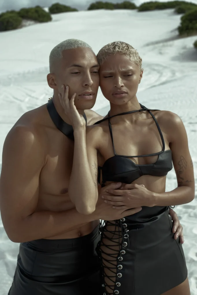 Fashion couple with platinum blonde hair in black leather attire embracing against white sand dunes
