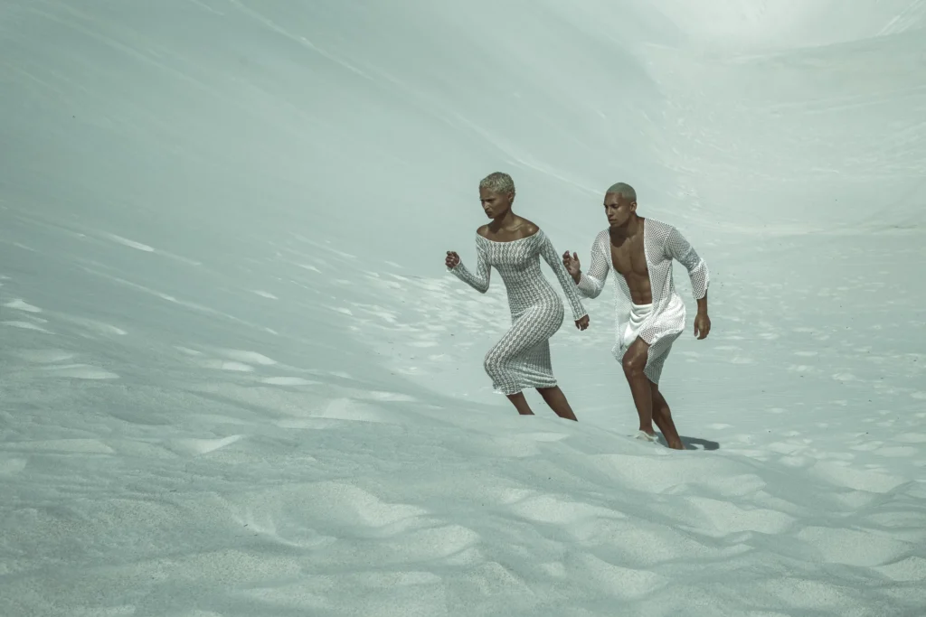 Two models in white knit garments running through pale sand dunes, ethereal fashion editorial