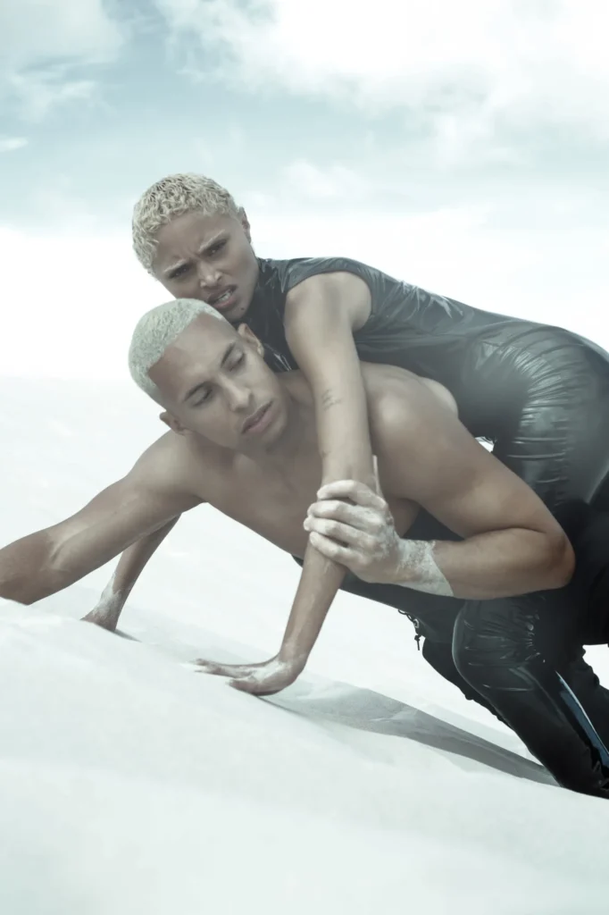 Two blonde models in black leather fashion editorial pose on white sand dunes, dramatic styling