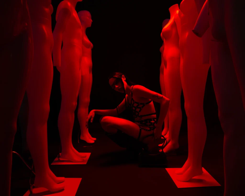 Woman crouching among mannequins in dramatic red lighting, edgy fashion editorial portrait