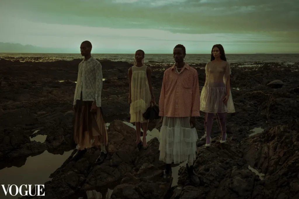 Four models in sheer layered garments standing on dramatic rocky coastline at dusk, editorial fashion shoot