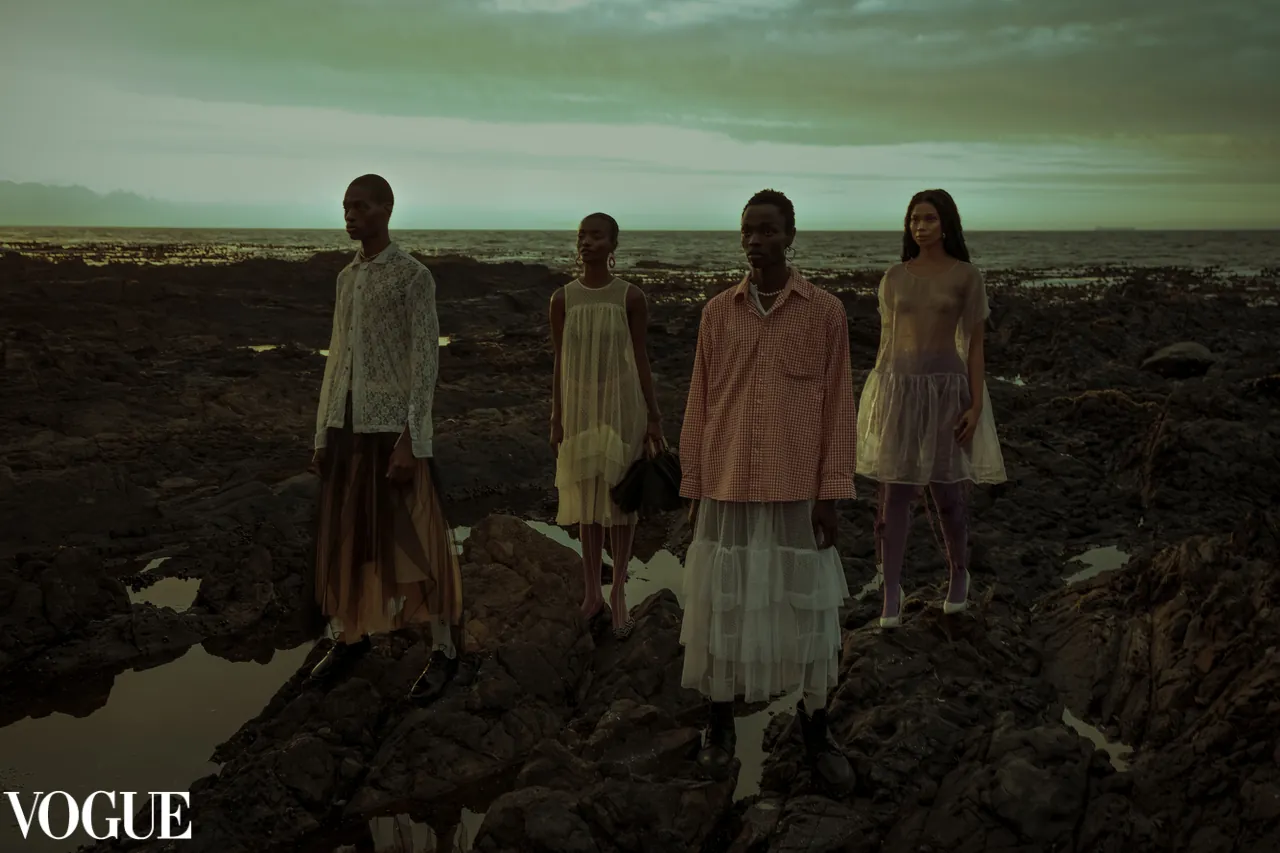 Four models in sheer layered garments standing on dramatic rocky coastline at dusk, editorial fashion shoot