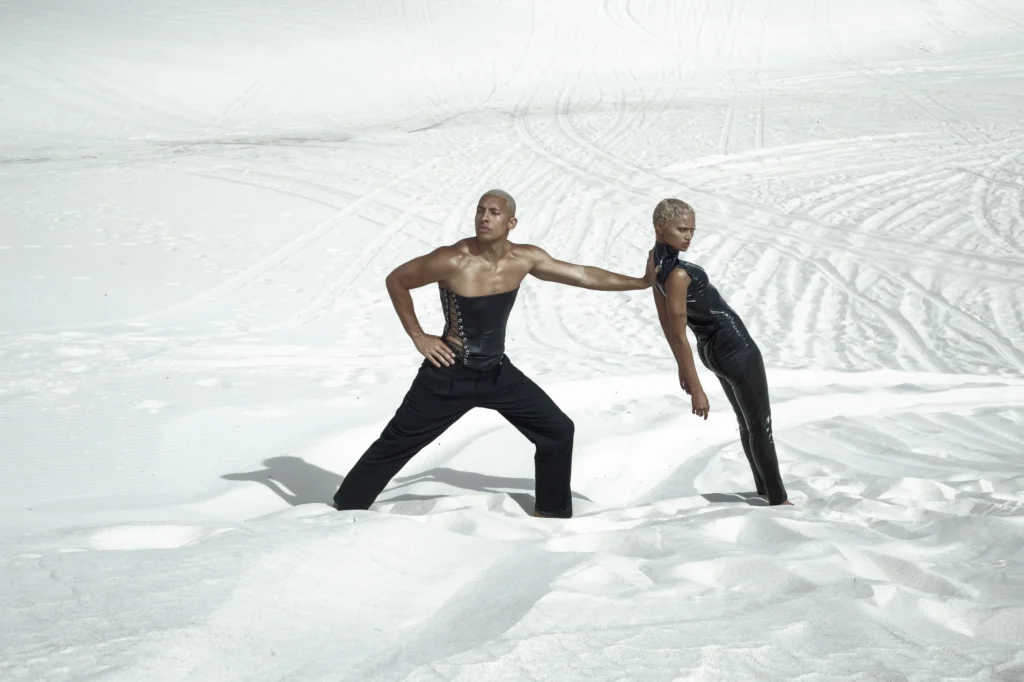 Two models in black avant-garde outfits posing dramatically on white sand dunes, fashion editorial